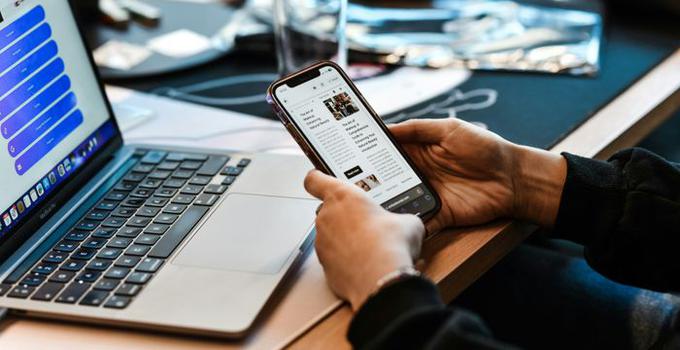 Dator på ett skrivbord samt en mobiltelefon i en persons hand. 