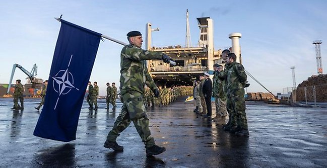 Militär som bär en Nato-flagga framför en båt. 