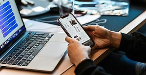 Dator på ett skrivbord samt en mobiltelefon i en persons hand. 